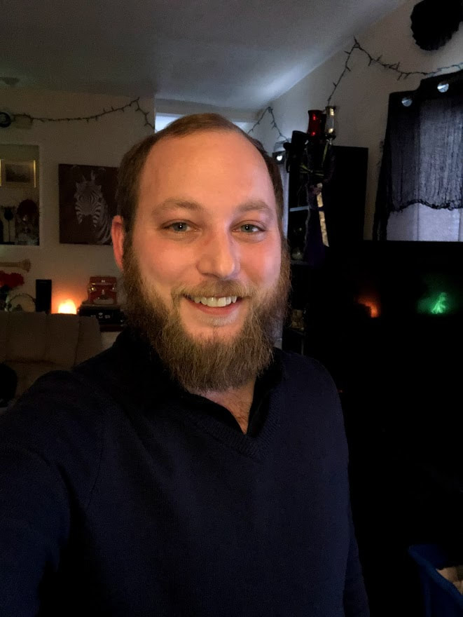 Dominick Hill. White male with a toothy smile taking a “selfie” in his friend’s living room. He has fair skin, blue eyes, brown hair (thinning on the top), and a 3 inch dirty blonde/light brown beard. He is wearing a blue sweater with a black collared shirt underneath. In the background are Christmas lights hung from the ceiling, although not turned on. To the right of Dominick, a TV is in the background reflecting an image of a dinosaur light outside of the view of the picture. There is a window behind the TV bookshelf next to that. To the left is a painting of a zebra’s head on the wall behind him and a couch. There is a salt lamp behind the couch, along with other unclear items.