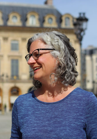 A white female with black-rimmed glasses and curly, salt and pepper hair is wearing a blue top. She is outside, turned to her right and laughing. Old buildings of Paris and a black lamp post are in the distant background.