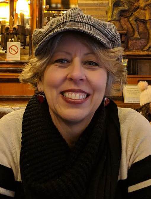 Female, caucasian, short wavy blonde hair with blue eyes, wearing a plaid hat, black and white turtle neck sweater with red heart earrings smiling and sitting in a restaurant with bar behind her.